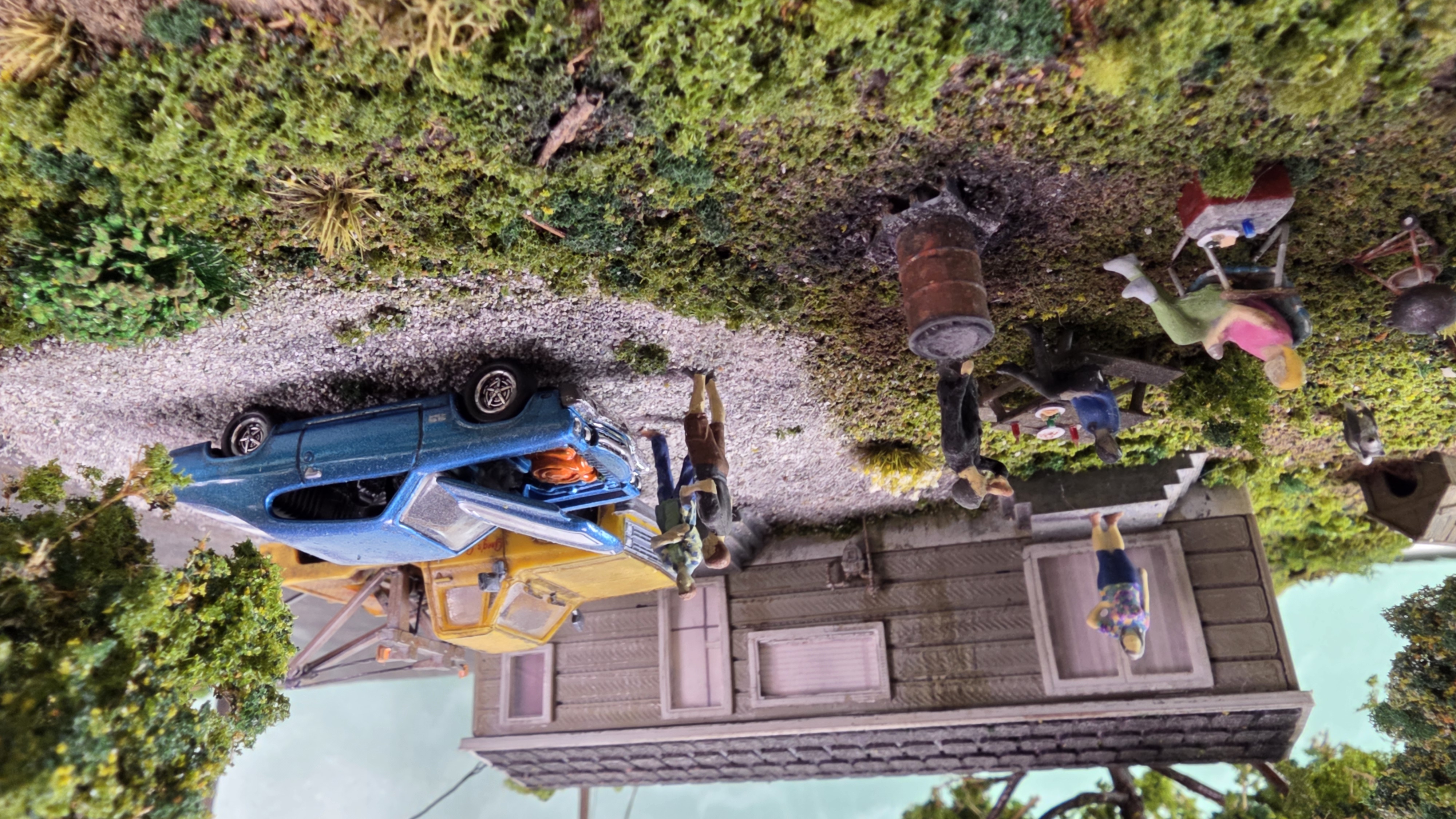 Daytime cookout in the side yard — figures gathered around the burn barrel and picnic table beside the blue car