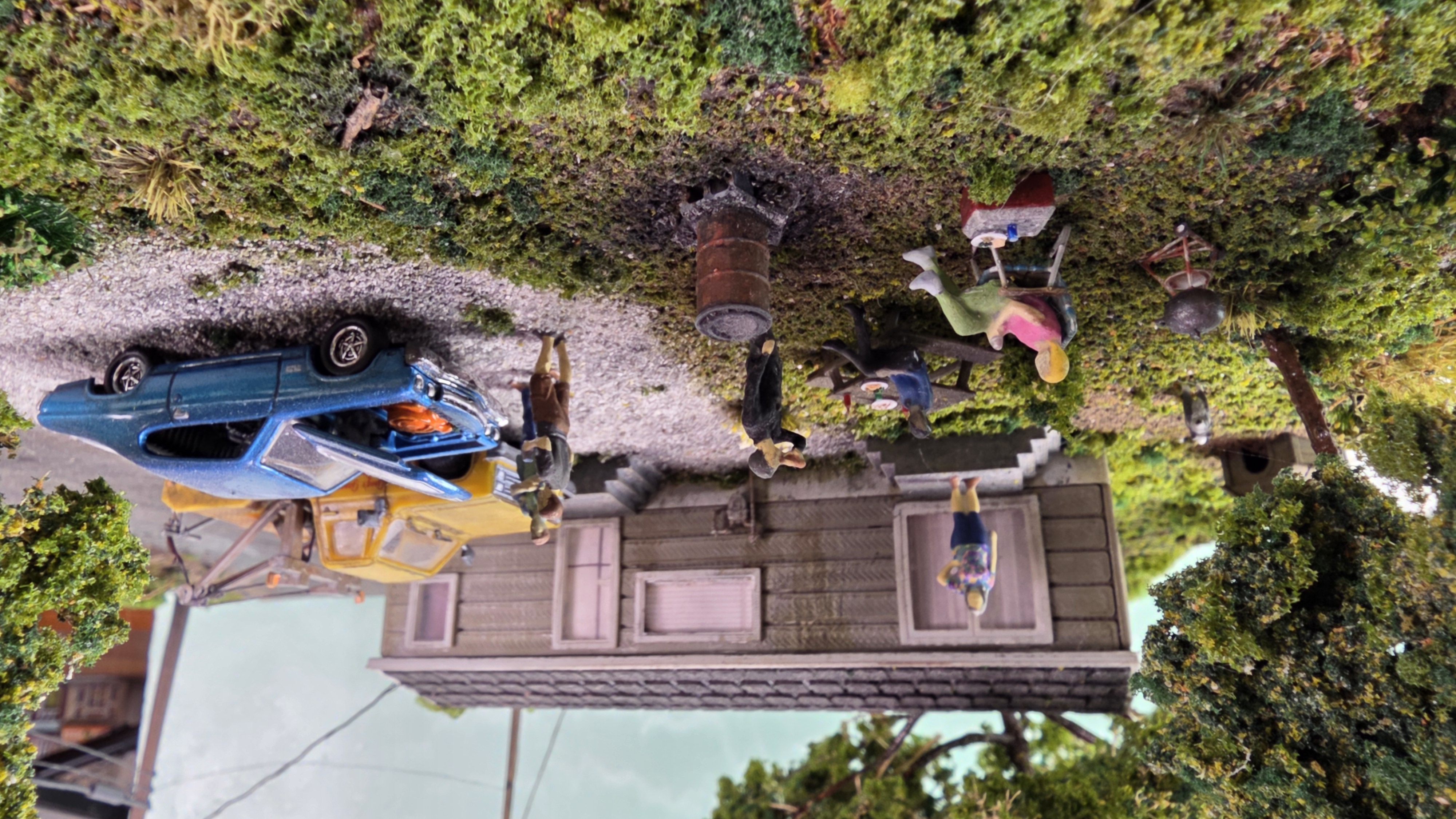 Daytime backyard cookout beside the trailer — figures around the picnic table, grill, and burn barrel with the blue car and tow truck